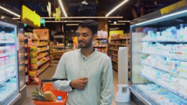 笑顔のインドの男は スーパーマーケットで彼のトロリーと通路を歩く — ストック動画