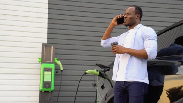 Чорний Чоловік Розмовляє Телефону Каву Заряджаючи Розкішний Електромобіль Вокзалі — стокове відео