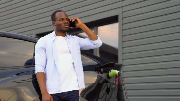 Czarny Mężczyzna Rozmawia Przez Telefon Podczas Ładowania Luksusowego Samochodu Elektrycznego — Wideo stockowe