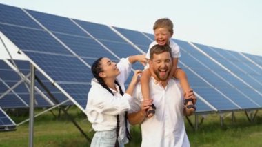 Üç kişilik genç bir aile fotovoltaik güneş panelinin yanında çömelmiş gülümsüyor ve parlak gelecek kavramına bakıyor.