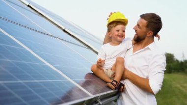 Güneş panelinin yanında şapkalı baba-oğul mühendislik konsepti. Baba oğlunu kollarında tutarken büyük bir güneş panelinin önünde ona yenilenebilir enerjinin temellerini gösteriyor.