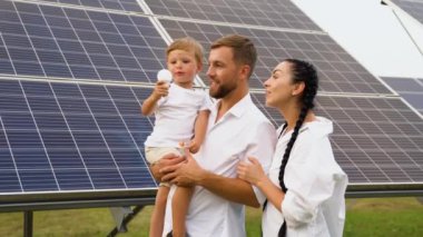 Güneş panelleri yakınında ampullü bir aile. Çocuk elinde bir ampul tutarken ebeveynleri onlara gururla ve hayranlıkla bakıyor..
