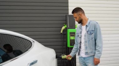 Bir adam istasyonda şarjlı elektrikli aracın fişini çekiyor. Yeşil enerji, çevre dostu yakıt.