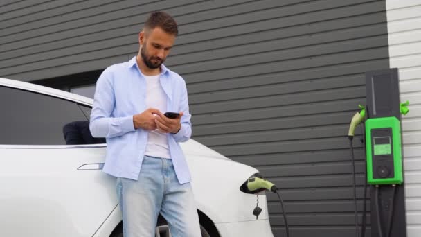 バッテリー充電プロセスの完了を待っている電気自動車の近くにスマートフォンを持つカジュアル男 — ストック動画