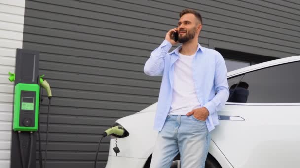 電気自動車の充電ステーションで車を充電しながら電話する男 — ストック動画
