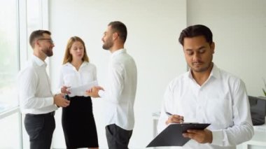 Hintli iş adamı kameraya bakıyor ve takım toplantısında bir ofiste duruyor..