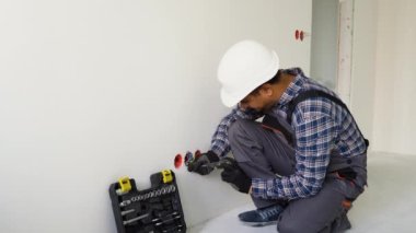 Hintli elektrikçi, prizler ve anahtarlar yerleştiriyor. Profesyonel işçi tulumu ve elektrikli alet..