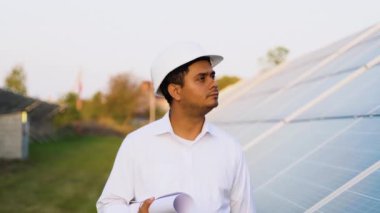 Güneş enerjisi santralindeki fotovoltaik panelleri inceleyen koruyucu miğfer mühendisi..
