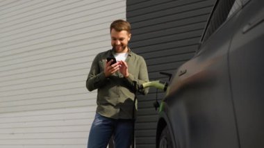 Elektrikli arabanın yanında duran bir adam şarj oluyor ve akıllı telefondan zaman ayarları yapıyor.