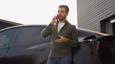 Gündelik kıyafetlerle gülümseyen beyaz adam, yeni lüks elektrikli arabasına yaslanırken konuşan telefonu takıyor, bir arabayı şarj ettiği için şarj istasyonunda duruyor.