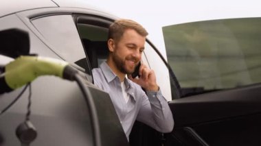 Şarj istasyonunda lüks elektrikli araba şarj ederken telefonda konuşan bir adam..