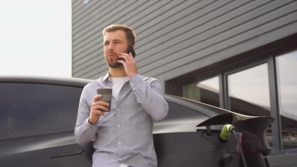 ハンサムな男が電話で話し 駅で高級電気自動車を充電しながらコーヒーを飲む — ストック動画