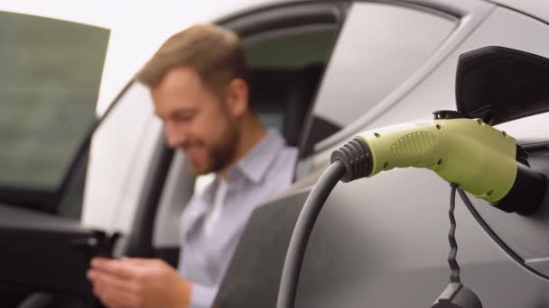 Homme Recharge Véhicule Électrique Avec Câble Regardant App Sur Téléphone — Video