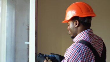 Siyah tamirci beyaz PVC plastik pencereyi içeri ayarlıyor. Afrikalı bir işçi pencereyi onarmak için tornavida kullanıyor. ev ödevi.