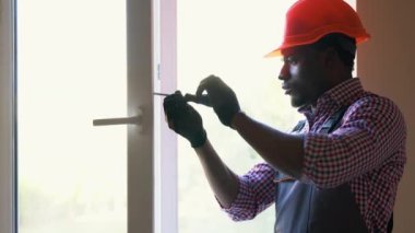 Siyah tamirci beyaz PVC plastik pencereyi içeri ayarlıyor. Afrikalı bir işçi pencereyi onarmak için tornavida kullanıyor. ev ödevi.