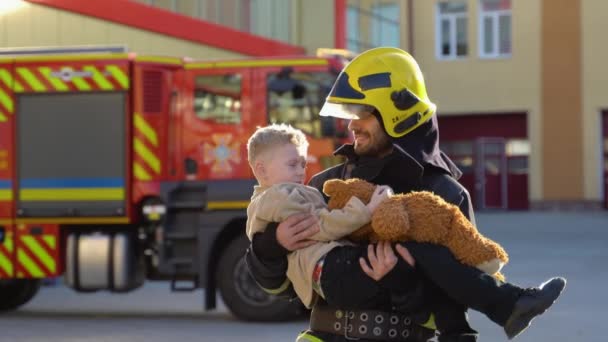 Portret Odważnego Strażaka Trzyma Uratowanego Chłopca Ramionach Stojącego Obok Wozu — Wideo stockowe