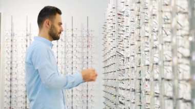 Bir adam optik dükkanında aynaya bakarak gözlük seçer..