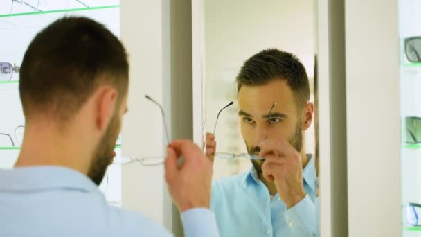 Ένας Άντρας Διαλέγει Γυαλιά Στο Κατάστημα Οπτικών Κοιτάζοντας Τον Καθρέφτη — Αρχείο Βίντεο