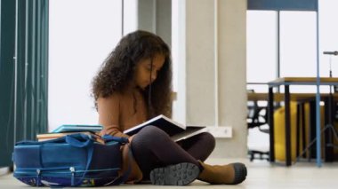 Siyah liseli kız sınıfta yerde oturmuş kitap okuyor..
