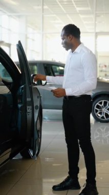 Gülümseyen, dost canlısı araba satıcısı araba kuaföründe açık kapı ve yeni araba gösteriyor.