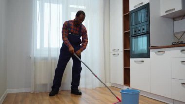 Üniformalı profesyonel bir siyah temizlikçi. Mutfakta paspas çubuğu ve kovayla yerleri yıkıyor..