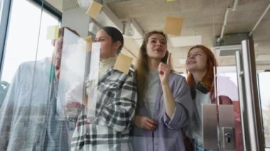 Genç, mutlu, profesyonel, yaratıcı grup öğrencileri beyin fırtınası yaparak modern ofis masasında başlangıç projesini tartışıyorlar cam üzerine yapışkan notlara bakıyorlar..