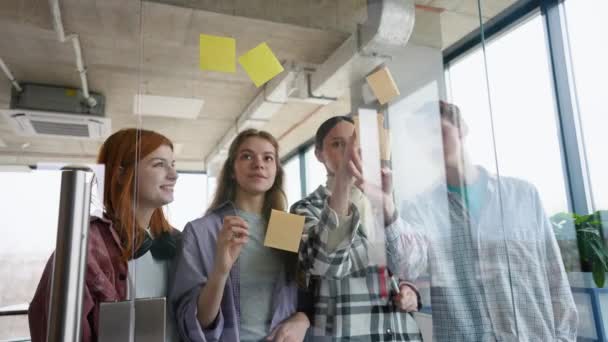 一组学生在头脑风暴时一起工作 站在玻璃墙后面 拿着色彩斑斓的粘纸片 — 图库视频影像