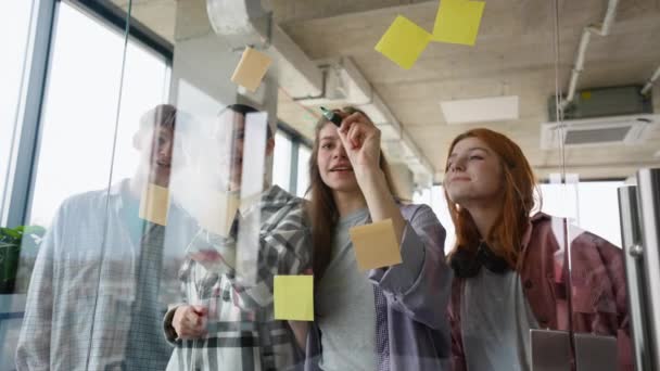 研究計画を議論し 粘着性のあるカラフルなペーパーでガラスの壁の後ろに立っている学生のグループ — ストック動画
