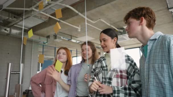 ブレインストーミング中に一緒に作業しながら 粘着性のあるカラフルなペーパーでガラスの壁の後ろに立っている学生のグループ — ストック動画