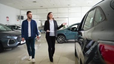 Kadın araba satıcısı galeride müşteriye yeni araba gösteriyor. Genç adam galeride araba seçerken kadın satış müdürüyle konuşuyor.