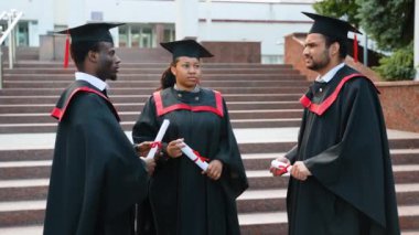 Kep ve cübbe giymiş üniversite mezunları konuşuyor. Eğitim, mezuniyet ve halk konsepti - bir grup mutlu uluslararası öğrenci önlük ve açık havada