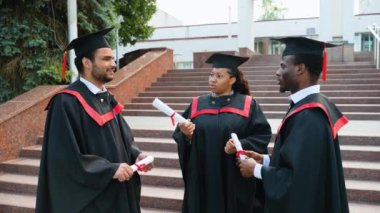 Kep ve cübbe giymiş üniversite mezunları konuşuyor. Eğitim, mezuniyet ve halk konsepti - bir grup mutlu uluslararası öğrenci önlük ve açık havada