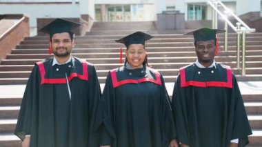 Üniversite kampüsünde poz veren mezuniyet elbisesi giymiş genç öğrenciler. Eğitim, mezuniyet ve halk konsepti - bir grup mutlu uluslararası öğrenci önlük ve açık havada