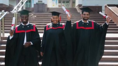 Mezuniyet elbisesi giymiş üç üniversite mezunu diplomayla kampüs boyunca yürüyorlar. Çok ırklı çeşitli mezunların kampüste diplomaları var. Mezuniyetleri mutlu geçiyor.