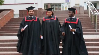 Üniversite kampüsünde yürüyen ve konuşan üç üniversite mezunu. Çok ırklı çeşitli mezunların kampüste diplomaları var. Mezuniyetleri mutlu geçiyor.