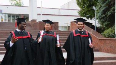 Üniversite kampüsünde yürüyen ve konuşan üç üniversite mezunu. Çok ırklı çeşitli mezunların kampüste diplomaları var. Mezuniyetleri mutlu geçiyor.