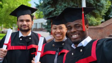 Mezuniyet elbisesi giymiş, özçekim yapan, gülümseyen ve mezuniyeti kutlayan üç farklı ırktan mutlu öğrenci. Mutlu anları yakalıyorum.