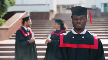 Mezuniyet elbisesi ve şapkasıyla yüzünde kocaman bir gülümsemeyle kameraya doğru poz veren Afro-Amerikan mezunu. Eğitim, mezuniyet ve halk konsepti - manto ve şapka giyen bir grup uluslararası öğrenci