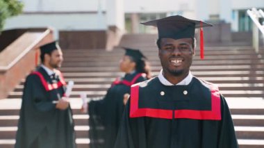 Üniversite kampüsündeki mezuniyet gününde gülümseyen genç bir adam çok ırklı bir arkadaş grubuyla duruyor. Eğitim, nitelik ve önlük konsepti