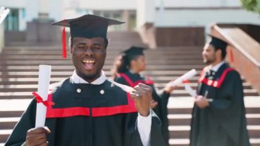Üniversite kampüsündeki mezuniyet gününde Afrikalı gülümseyen bir öğrenci çok ırklı bir arkadaş grubuyla duruyor. Eğitim, mezuniyet ve halk konsepti - bir grup uluslararası öğrenci manto ve