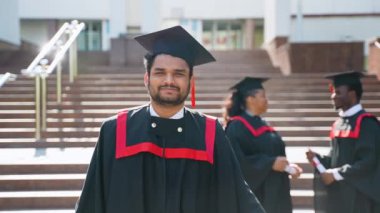 Mezuniyet kostümlü Hintli öğrenci diplomasını gösteriyor ve kameraya gülümsüyor. Eğitim, mezuniyet ve halk konsepti - dış mekan ve şapkalar içinde uluslararası öğrenciler grubu