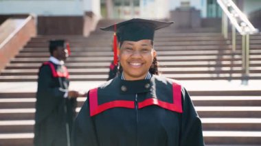 Afro-Amerikalı kadın mezuniyet elbisesi ve şapkasıyla kameraya doğru poz veriyor. Eğitim, mezuniyet ve halk konsepti - bir grup uluslararası öğrenci manto içinde