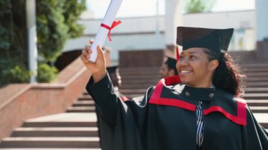 Mezuniyet gününde üniversite kampüsünde diploması olan Afrikalı Amerikalı bir kız öğrenci. Eğitim, mezuniyet ve halk konsepti - dış mekan ve şapkalar içinde uluslararası öğrenciler grubu