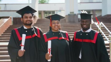 Üniversite kampüsünde mezuniyet elbisesi giymiş bir grup genç öğrenci sarılıyor. Eğitim, mezuniyet ve halk konsepti - dış mekan ve şapkalar içinde uluslararası öğrenciler grubu