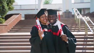 Mezuniyet elbiseleri ve şapkalarıyla üniversite kampüsünde poz veren mutlu Afro-Amerikan çift öğrencileri. Mezunlar mezuniyetin tadını çıkarıyor ve kutluyor, yakın plan portre