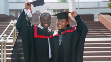 Mezuniyet elbiseleri ve şapkalarıyla üniversite kampüsünde poz veren mutlu Afro-Amerikan çift öğrencileri. Mezunlar mezuniyetin tadını çıkarıyorlar.
