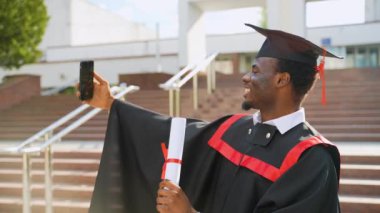 Video konferansındaki öğrenci mezuniyeti ailelerin diplomasını gösteriyor, mezuniyeti kutluyor. Ailesi için bir selfie videosu çekmek için kamerayı kullanan Afro-Amerikan mezunu bir şeyler konuşuyor ve el ele tutuşuyor.