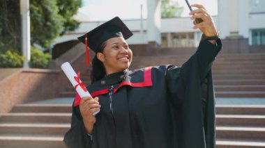 Videodaki kız öğrenci mezuniyeti ailelerin diplomasını gösteriyor, mezuniyeti kutluyor. Afrikalı Amerikan mezunu. Kamerayla ailesine selfie videosu çekiyor. Bir şeyler konuşuyor ve elinde tutuyor.