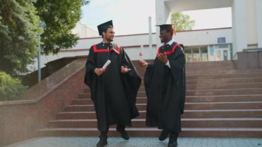 Mezuniyet mantoları içinde üniversite kampüsünde yürüyüp konuşan iki çok ırklı üniversite mezunu. Eğitim, mezuniyet ve halk konsepti - iki uluslararası önlüklü ve açık hava şapkalı öğrenci