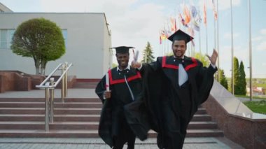 Afrikalı Amerikalı ve Hintli mezunlar mezuniyeti kutluyor, mezunlar kampüste koşuyor. Eğitim, mezuniyet ve halk konsepti - iki uluslararası önlüklü ve açık hava şapkalı öğrenci